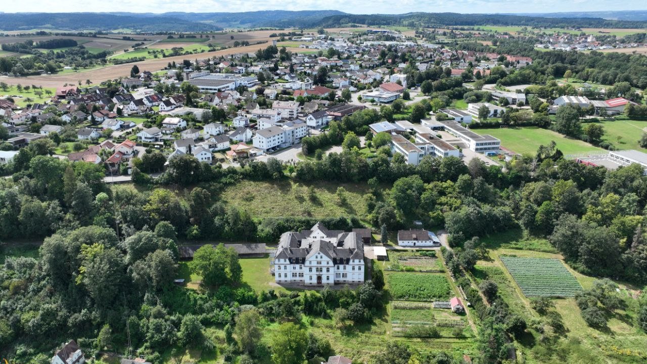 Luftaufnahme: die Vorderseite des Missionshauses zwischen Bäumen und Gärten, dahinter der Ort